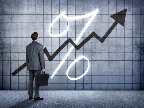 A businessman, standing while holding a briefcase, looks up at a chart on a wall that indicates interest rates are rising.